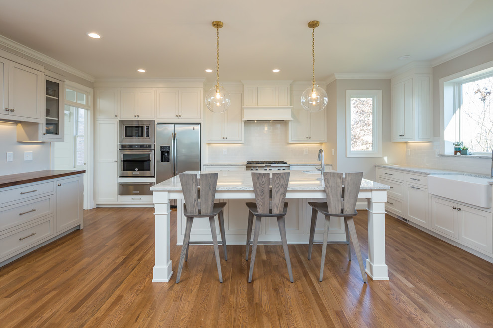 Inspiration for a large transitional u-shaped eat-in kitchen in Raleigh with a farmhouse sink, shaker cabinets, white cabinets, white splashback, stainless steel appliances, medium hardwood floors, with island, brown floor, quartzite benchtops, subway tile splashback and white benchtop.