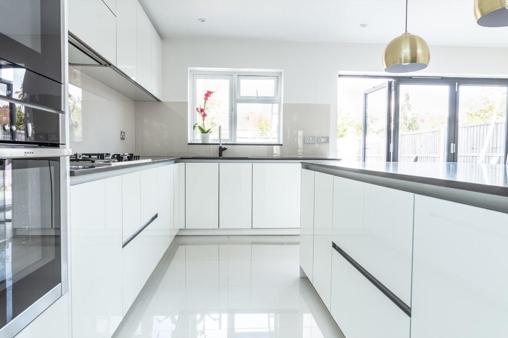 Photo of a contemporary l-shaped kitchen in London with a drop-in sink, flat-panel cabinets, white cabinets, grey splashback, glass sheet splashback, panelled appliances, with island, white floor and grey benchtop.