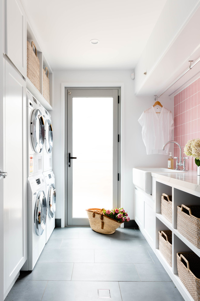 Photo of a mid-sized beach style galley dedicated laundry room in Brisbane with a farmhouse sink, open cabinets, white cabinets, a stacked washer and dryer, grey floor, white walls and white benchtop.