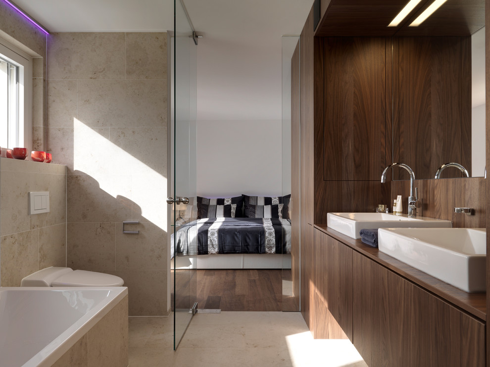 This is an example of a mid-sized bathroom in Stuttgart with flat-panel cabinets, medium wood cabinets, a drop-in tub, beige tile, stone tile, limestone floors, a vessel sink and wood benchtops.