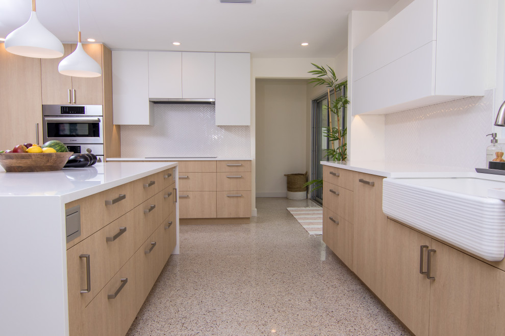 This is an example of a large modern l-shaped open plan kitchen in Tampa with a farmhouse sink, flat-panel cabinets, light wood cabinets, solid surface benchtops, white splashback, stainless steel appliances, terrazzo floors, with island, multi-coloured floor and white benchtop.