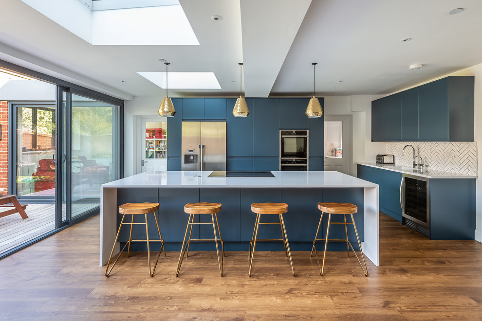 Large modern open plan kitchen in London with a drop-in sink, flat-panel cabinets, blue cabinets, quartzite benchtops, white splashback, subway tile splashback, stainless steel appliances, vinyl floors, with island and white benchtop.