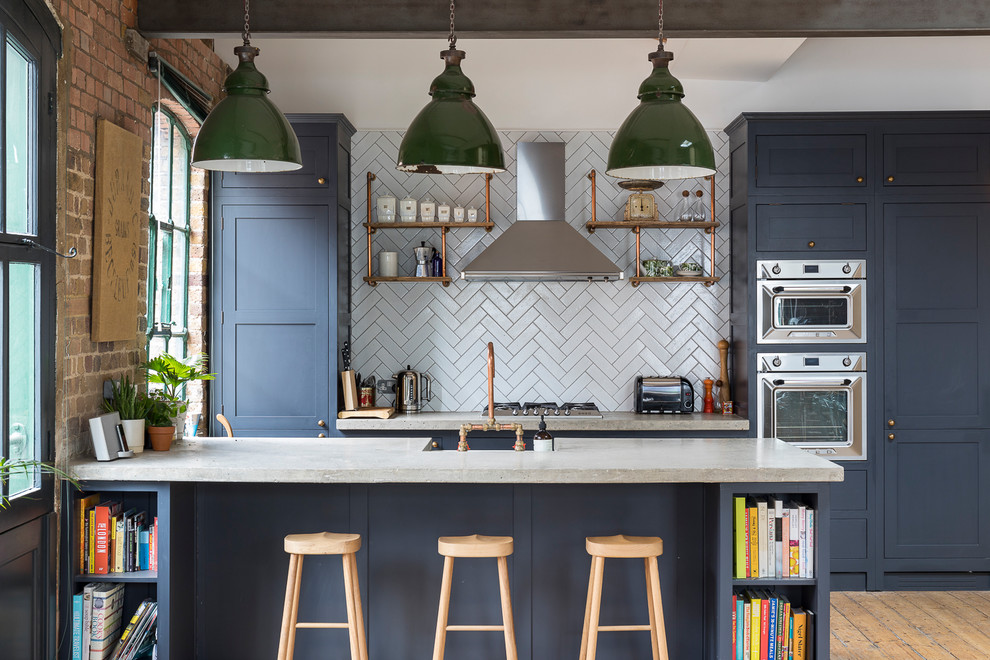 Photo of a large industrial galley kitchen in London with shaker cabinets, blue cabinets, grey splashback, stainless steel appliances, medium hardwood floors, a peninsula and brown floor.