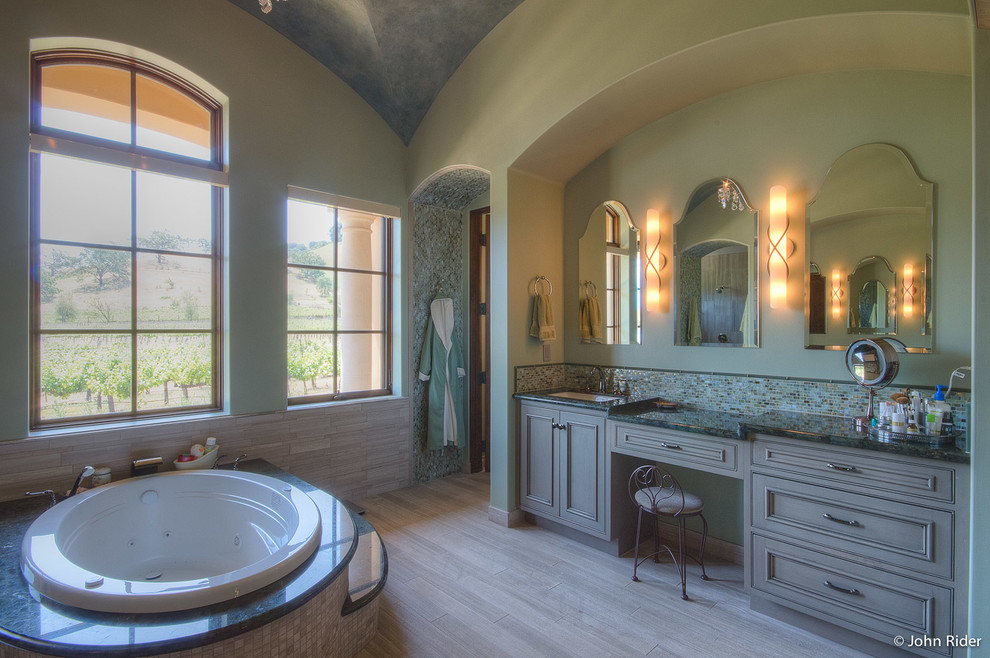 Photo of a large mediterranean master bathroom in San Francisco with an undermount sink, beaded inset cabinets, grey cabinets, granite benchtops, a drop-in tub, green tile, glass sheet wall, green walls and porcelain floors.
