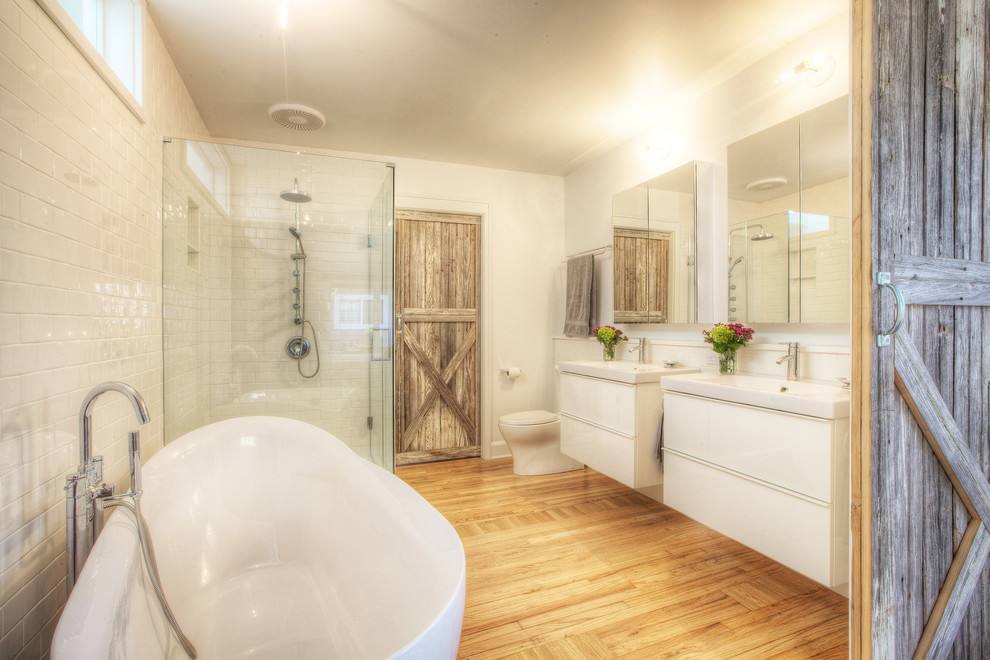 This is an example of a mid-sized country master bathroom in Indianapolis with a console sink, flat-panel cabinets, white cabinets, solid surface benchtops, a freestanding tub, a corner shower, a two-piece toilet, white tile, subway tile, white walls and light hardwood floors.