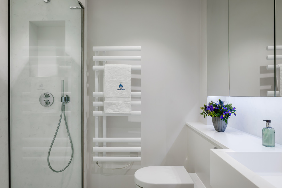 This is an example of a mid-sized contemporary master bathroom in London with flat-panel cabinets, white cabinets, a wall-mount toilet, porcelain floors, an integrated sink, solid surface benchtops, a sliding shower screen and a built-in vanity.