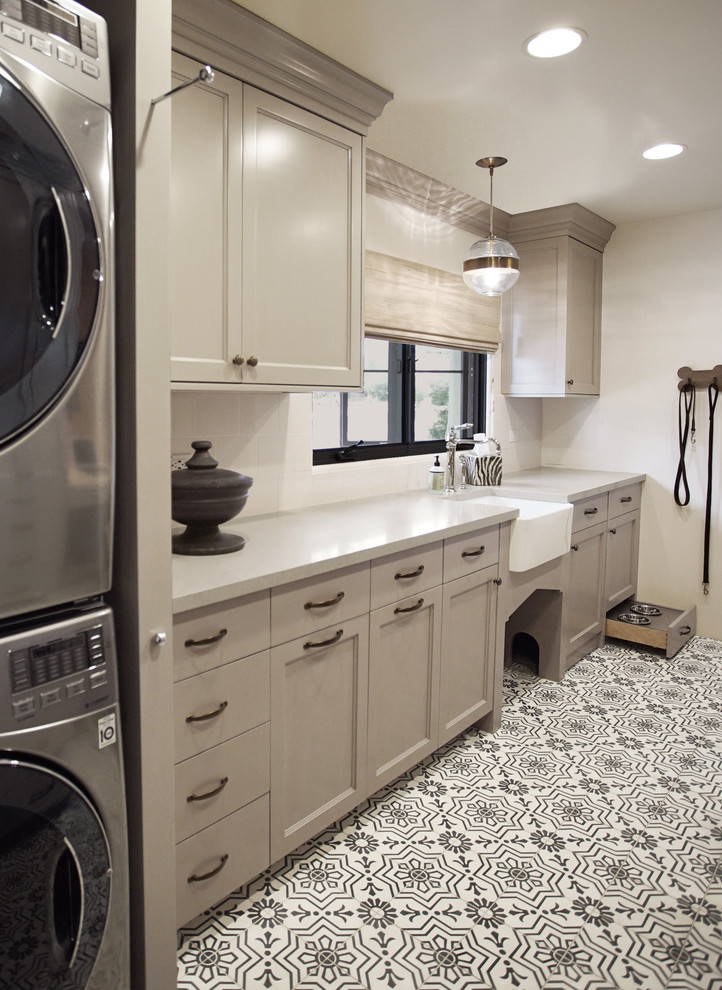 Example of a mid-sized transitional galley multicolored floor dedicated laundry room design in Phoenix with a farmhouse sink, recessed-panel cabinets, gray cabinets, quartz countertops, white backsplash, subway tile backsplash, white walls, a stacked washer/dryer and gray countertops