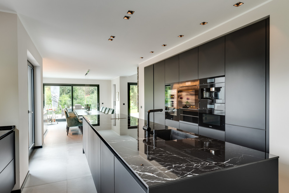 Example of a huge trendy galley ceramic tile and gray floor open concept kitchen design in Hamburg with flat-panel cabinets, black cabinets, quartz countertops, black appliances, an island, black countertops and an undermount sink