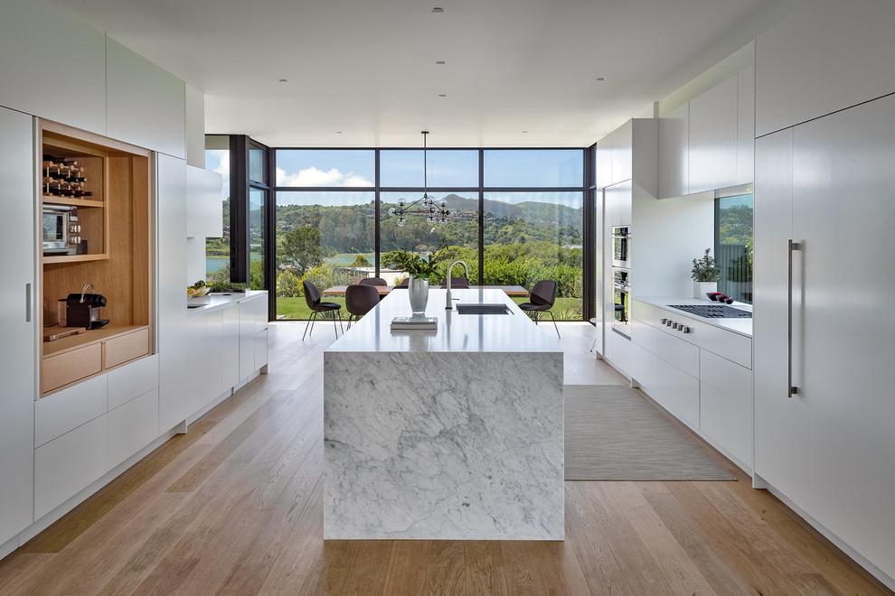 Large modern eat-in kitchen in San Francisco with an undermount sink, flat-panel cabinets, white cabinets, marble benchtops, panelled appliances, light hardwood floors, with island, white benchtop, window splashback and beige floor.