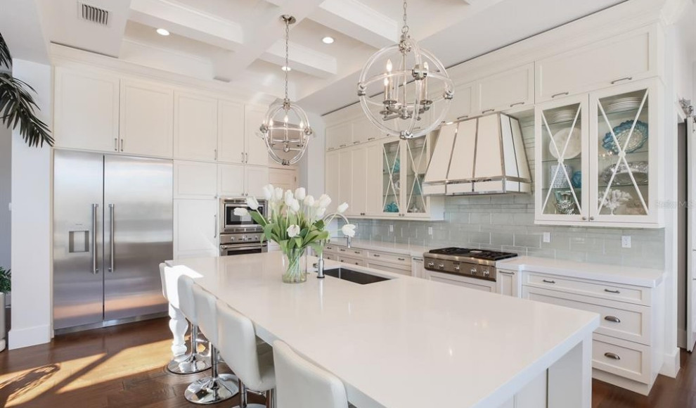 This is an example of an expansive contemporary l-shaped open plan kitchen in Tampa with a single-bowl sink, shaker cabinets, white cabinets, quartzite benchtops, white splashback, glass tile splashback, stainless steel appliances, light hardwood floors, with island, white benchtop and coffered.