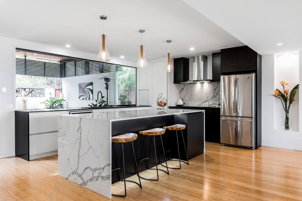 Photo of a large contemporary l-shaped open plan kitchen in Brisbane with a double-bowl sink, flat-panel cabinets, black cabinets, quartz benchtops, white splashback, stone slab splashback, stainless steel appliances, light hardwood floors and with island.