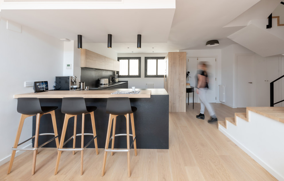 Scandinavian l-shaped open plan kitchen in Other with flat-panel cabinets, light wood cabinets, black splashback, light hardwood floors, a peninsula, beige floor and black benchtop.