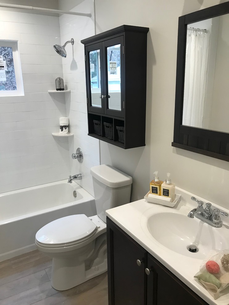 Inspiration for a transitional bathroom in New York with recessed-panel cabinets, black cabinets, an alcove tub, a shower/bathtub combo, a two-piece toilet, white tile, ceramic tile, white walls, medium hardwood floors, an integrated sink, solid surface benchtops, brown floor, a shower curtain and white benchtops.