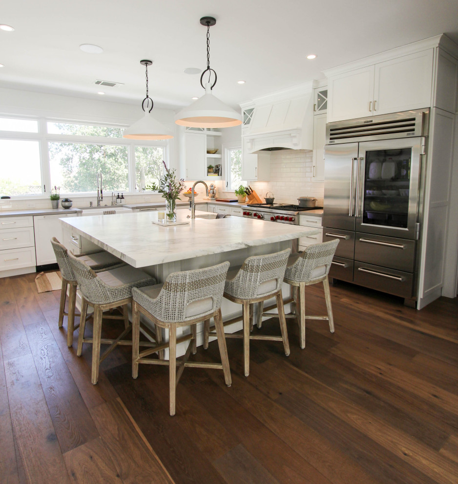 Large transitional u-shaped open plan kitchen in San Francisco with a farmhouse sink, shaker cabinets, white cabinets, quartz benchtops, white splashback, ceramic splashback, medium hardwood floors, with island, brown floor, white benchtop and stainless steel appliances.