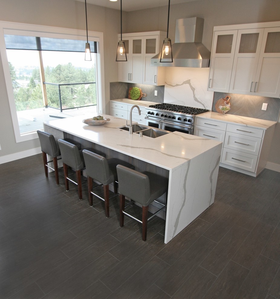 This is an example of a mid-sized transitional l-shaped open plan kitchen in Seattle with a farmhouse sink, shaker cabinets, white cabinets, quartz benchtops, grey splashback, porcelain splashback, stainless steel appliances, porcelain floors, with island, black floor and white benchtop.