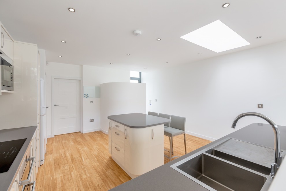 This is an example of a mid-sized contemporary l-shaped eat-in kitchen in Edinburgh with a double-bowl sink, flat-panel cabinets, beige cabinets, granite benchtops, grey splashback, stone slab splashback, panelled appliances, bamboo floors and with island.