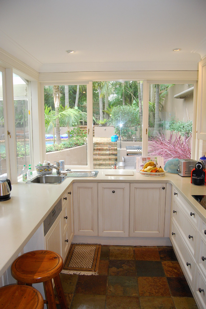 Photo of a mid-sized traditional u-shaped kitchen pantry in Sydney with a drop-in sink, shaker cabinets, distressed cabinets, solid surface benchtops, beige splashback, porcelain splashback, stainless steel appliances, slate floors and a peninsula.