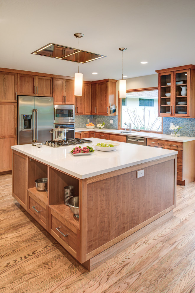 Mid-sized arts and crafts l-shaped open plan kitchen in San Francisco with an undermount sink, shaker cabinets, light wood cabinets, quartz benchtops, blue splashback, glass tile splashback, stainless steel appliances, light hardwood floors, with island and brown floor.