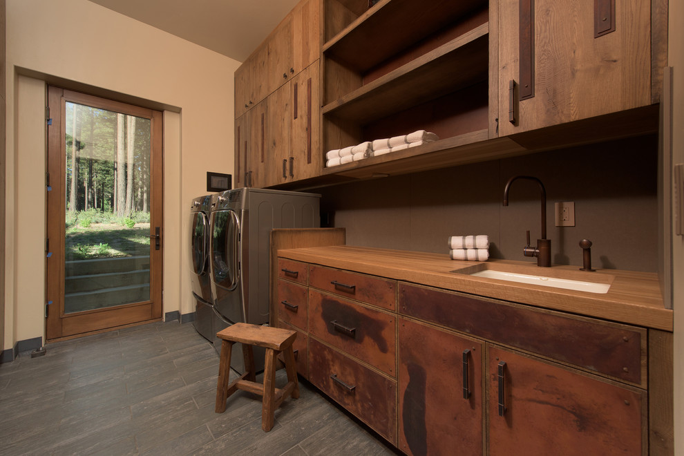 Inspiration for a large industrial single-wall dedicated laundry room in Other with an undermount sink, flat-panel cabinets, medium wood cabinets, wood benchtops, beige walls, slate floors and a side-by-side washer and dryer.