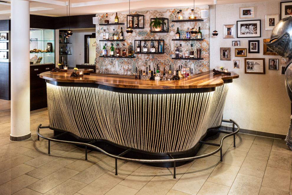 This is an example of a large industrial u-shaped seated home bar in Other with an integrated sink, flat-panel cabinets, black cabinets, wood benchtops, multi-coloured splashback, subway tile splashback, travertine floors, beige floor and brown benchtop.