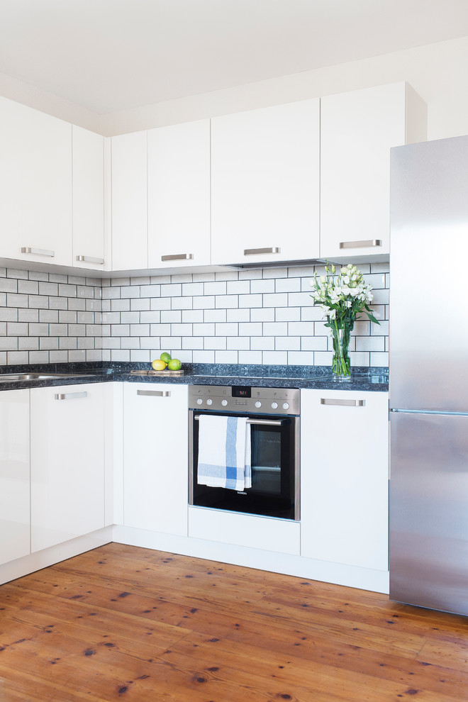 Inspiration for a small contemporary l-shaped separate kitchen in Berlin with white cabinets, granite benchtops, white splashback, subway tile splashback, stainless steel appliances, light hardwood floors, flat-panel cabinets and no island.