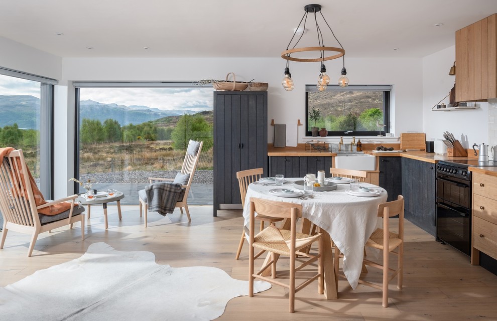 This is an example of a mid-sized scandinavian l-shaped open plan kitchen in Cornwall with a farmhouse sink, wood benchtops, white splashback, porcelain splashback, black appliances, light hardwood floors, beige floor, beige benchtop and grey cabinets.