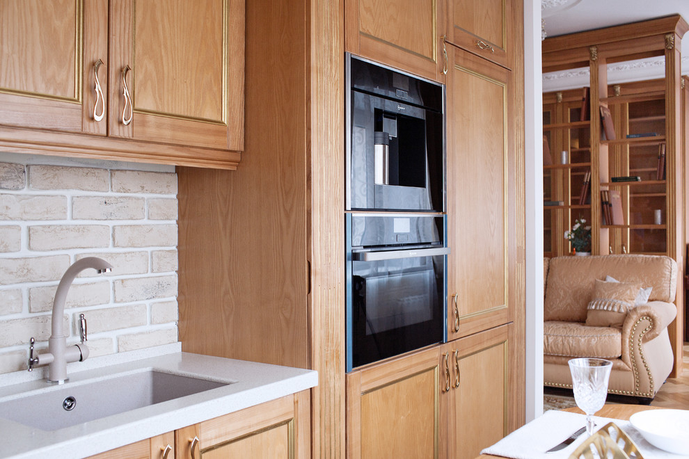This is an example of a small transitional l-shaped separate kitchen in Moscow with recessed-panel cabinets, solid surface benchtops, beige splashback, brick splashback, black appliances, no island, an undermount sink, light wood cabinets, white benchtop, porcelain floors and beige floor.