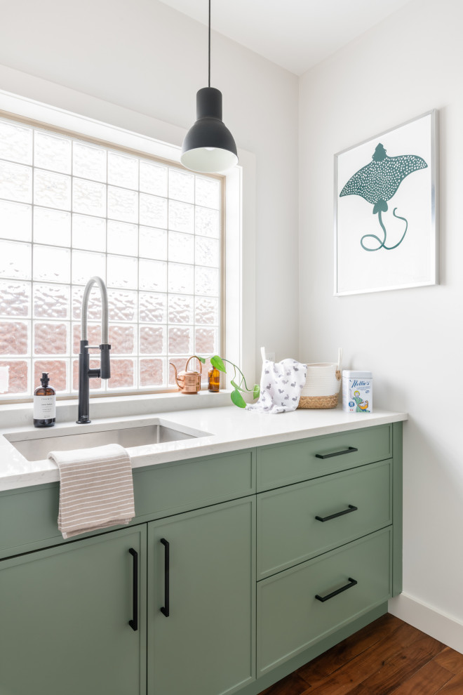Foto på en mellanstor vintage vita parallell tvättstuga, med en undermonterad diskho, skåp i shakerstil, gröna skåp, bänkskiva i kvarts, vitt stänkskydd, stänkskydd i keramik, mellanmörkt trägolv och brunt golv