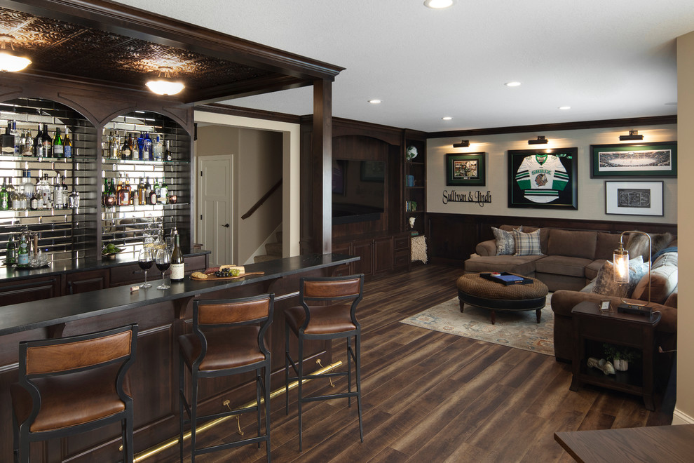 Mid-sized transitional galley seated home bar in Minneapolis with raised-panel cabinets, dark wood cabinets, soapstone benchtops, mirror splashback, vinyl floors, brown floor and black benchtop.