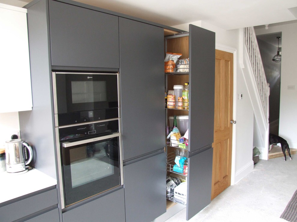 Inspiration for a mid-sized contemporary l-shaped eat-in kitchen in Dorset with a drop-in sink, flat-panel cabinets, grey cabinets, solid surface benchtops, white splashback, glass sheet splashback, black appliances, vinyl floors and no island.