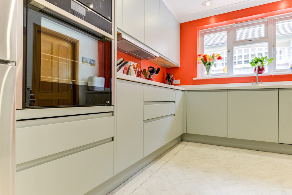 Photo of a medium sized nautical grey and white l-shaped enclosed kitchen in Sussex with a built-in sink, flat-panel cabinets, green cabinets, composite countertops, orange splashback, glass sheet splashback, black appliances, lino flooring, a breakfast bar, white floors, white worktops and feature lighting.