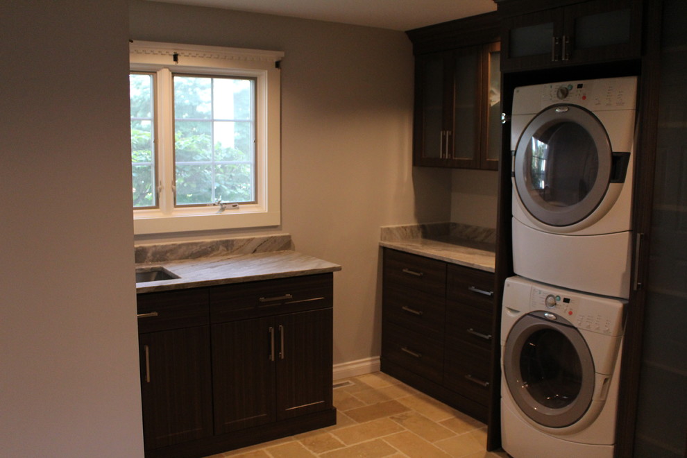 Photo of a small transitional l-shaped dedicated laundry room in Detroit with an undermount sink, shaker cabinets, dark wood cabinets, granite benchtops, grey walls, a stacked washer and dryer, beige floor, brown benchtop and ceramic floors.