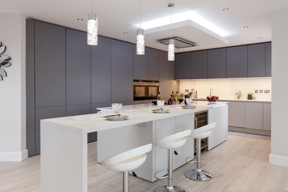 This is an example of a medium sized contemporary l-shaped kitchen in Dorset with flat-panel cabinets, grey cabinets, composite countertops, white splashback, integrated appliances, light hardwood flooring, an island and beige floors.