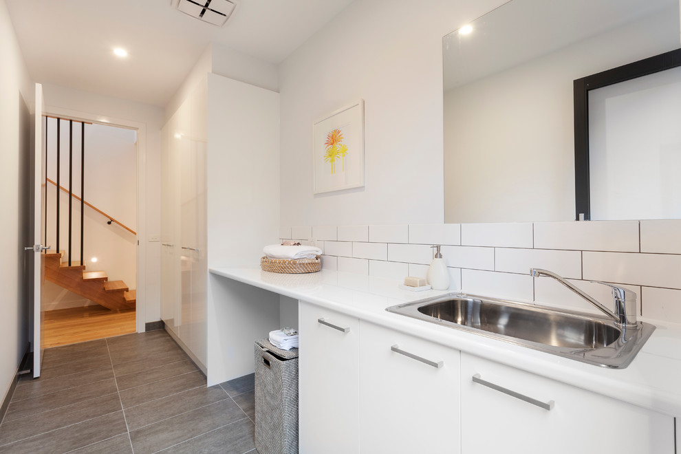 Mid-sized contemporary single-wall dedicated laundry room in Melbourne with a drop-in sink, flat-panel cabinets, white cabinets, solid surface benchtops, white walls, a concealed washer and dryer and grey floor.