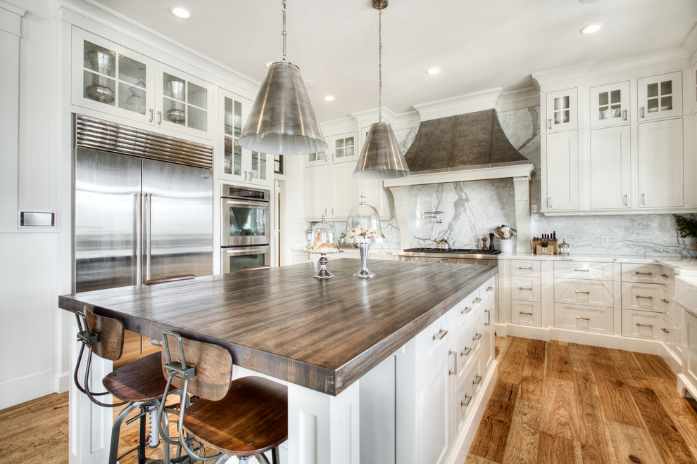 Photo of a large transitional u-shaped eat-in kitchen in Calgary with a farmhouse sink, shaker cabinets, white cabinets, marble benchtops, white splashback, stone slab splashback, stainless steel appliances, medium hardwood floors, with island and brown floor.