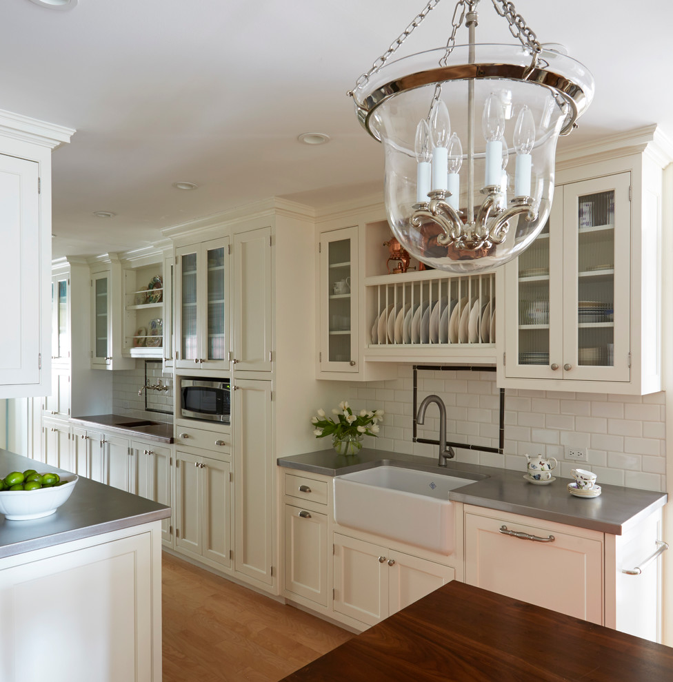 Inspiration for a large traditional galley separate kitchen in Chicago with a farmhouse sink, shaker cabinets, white cabinets, stainless steel benchtops, white splashback, subway tile splashback, panelled appliances, light hardwood floors and with island.