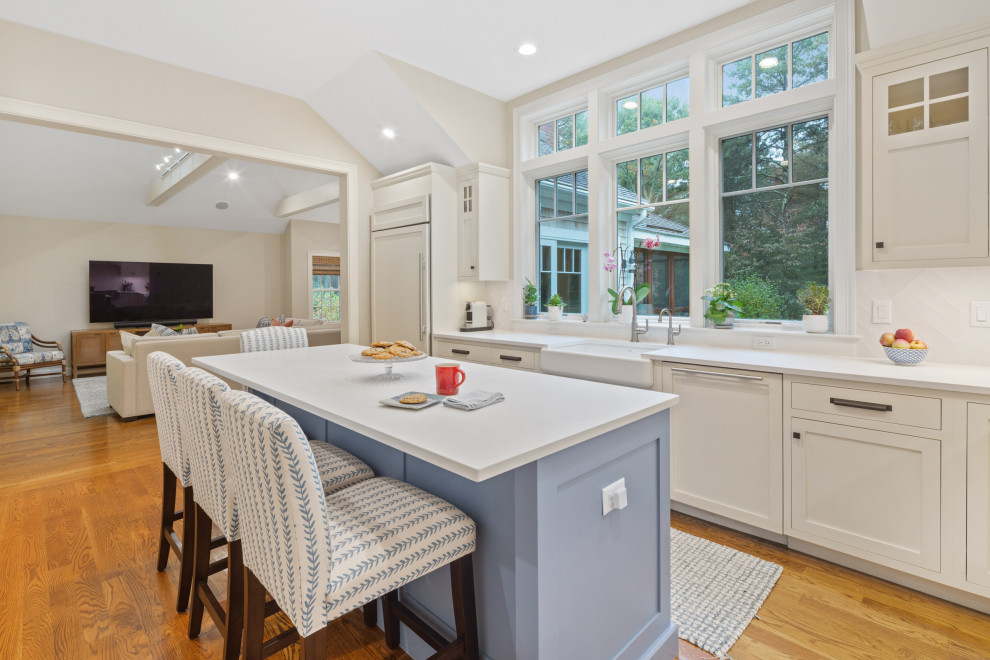 Kitchen pantry - large coastal l-shaped medium tone wood floor and brown floor kitchen pantry idea in Boston with a farmhouse sink, shaker cabinets, white cabinets, quartz countertops, white backsplash, ceramic backsplash, stainless steel appliances, an island and white countertops