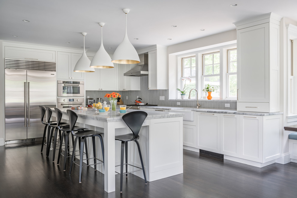 This is an example of a transitional l-shaped kitchen in Boston with a farmhouse sink, shaker cabinets, white cabinets, grey splashback, stainless steel appliances, dark hardwood floors, with island and grey benchtop.