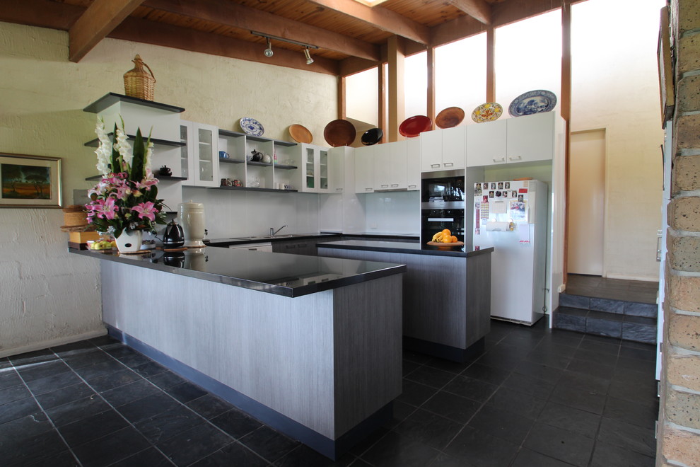 This is an example of a large contemporary u-shaped open plan kitchen in Melbourne with a drop-in sink, flat-panel cabinets, grey cabinets, granite benchtops, white splashback, glass sheet splashback, stainless steel appliances, slate floors and multiple islands.