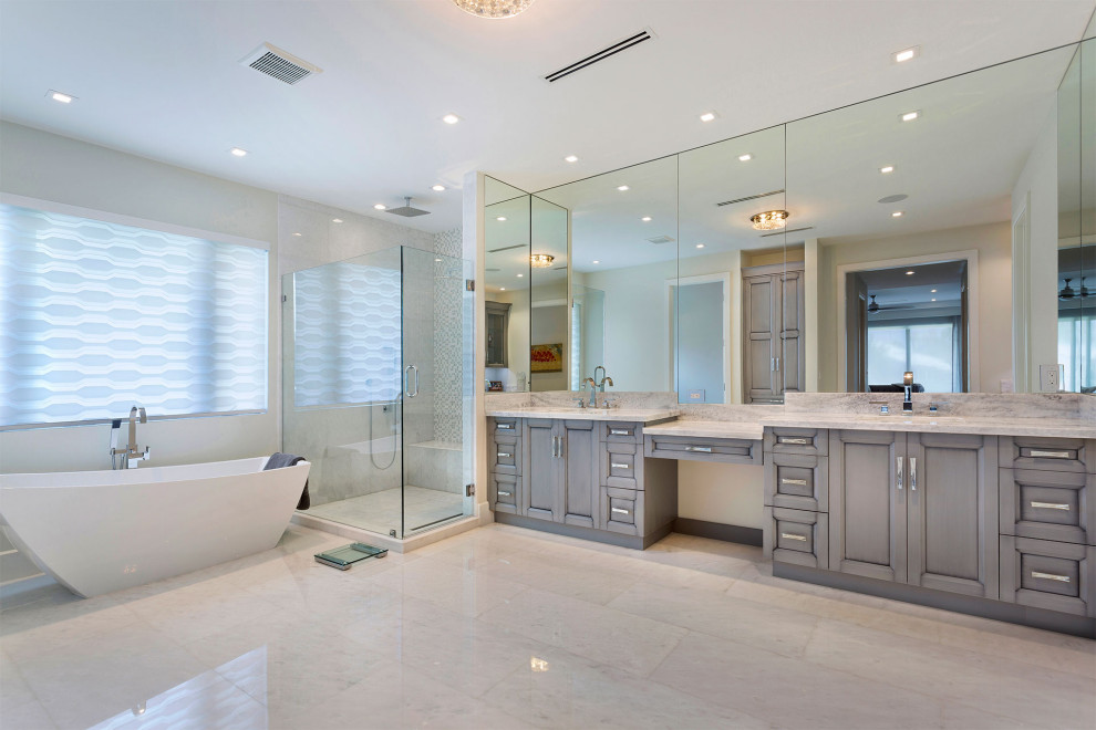 Large master bathroom in Miami with recessed-panel cabinets, grey cabinets, a freestanding tub, a corner shower, a one-piece toilet, white tile, pebble tile, beige walls, porcelain floors, an undermount sink, marble benchtops, beige floor, a hinged shower door and multi-coloured benchtops.