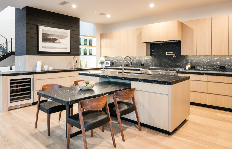 Contemporary l-shaped eat-in kitchen in Los Angeles with an undermount sink, flat-panel cabinets, light wood cabinets, black splashback, stainless steel appliances, light hardwood floors, with island, black benchtop, marble benchtops and marble splashback.