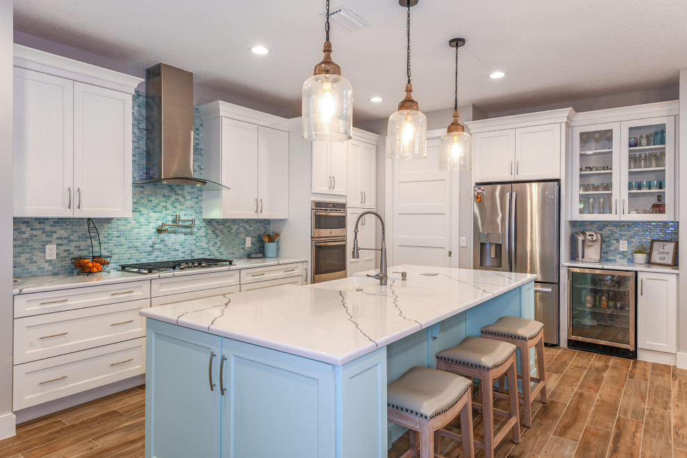 Photo of a mid-sized beach style l-shaped open plan kitchen in Orlando with a farmhouse sink, shaker cabinets, white cabinets, granite benchtops, multi-coloured splashback, glass tile splashback, stainless steel appliances, ceramic floors, with island, brown floor and white benchtop.
