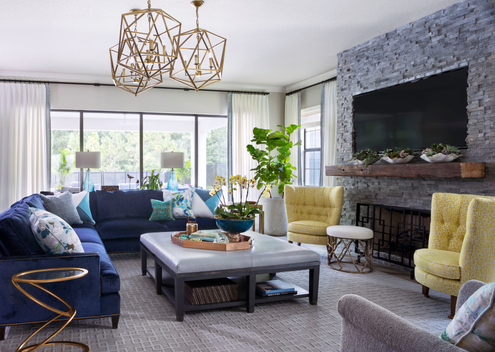 Colorful Great Room - This Great room is where the family spends a majority of their time.  A large navy blue velvet sectional is extra deep for hanging out or watching movies. We layered floral pillows, color blocked pillows, and a vintage rug fragment turned decorative pillow on the sectional.  The sunny yellow chairs flank the fireplace and an oversized custom gray leather cocktail ottoman does double duty as a coffee table and extra seating.  The large wood tray warms up the cool color palette.  A trio of openwork brass chandeliers are scaled for the large space.  We created a vertical element in the room with stacked gray stone and installed a reclaimed timber as a mantel.