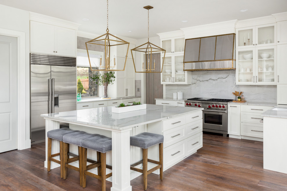 This is an example of a transitional u-shaped kitchen in DC Metro with a farmhouse sink, shaker cabinets, white cabinets, marble benchtops, grey splashback, marble splashback, stainless steel appliances, medium hardwood floors, with island, brown floor and grey benchtop.