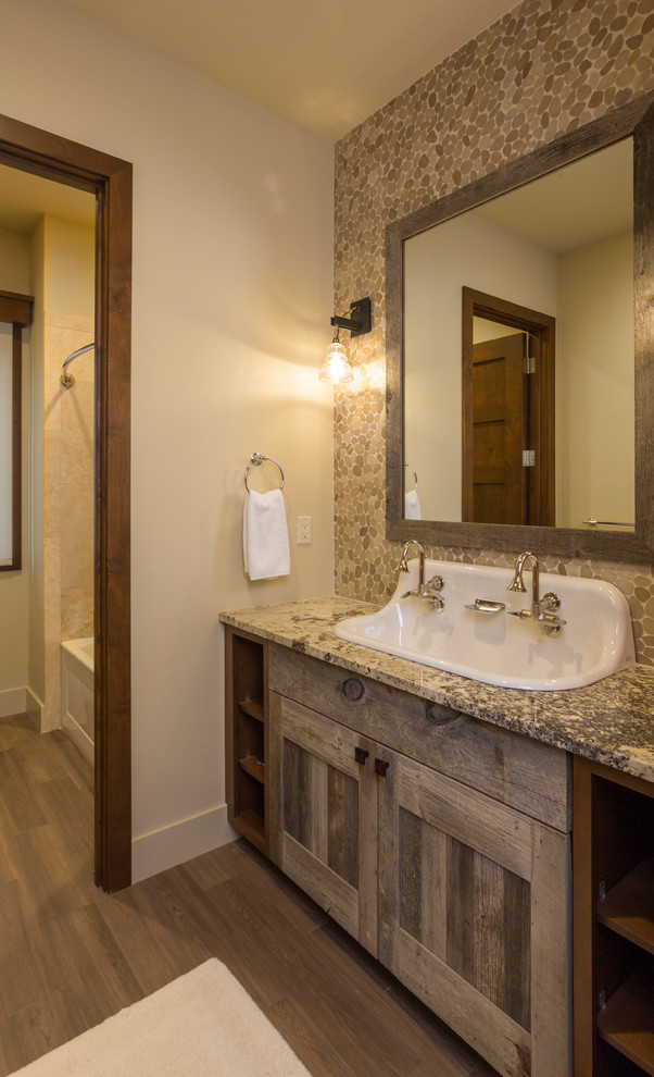 Photo of a mid-sized contemporary kids bathroom in Denver with recessed-panel cabinets, distressed cabinets, an alcove tub, a shower/bathtub combo, multi-coloured tile, pebble tile, beige walls, medium hardwood floors, a trough sink, granite benchtops, brown floor, a shower curtain and multi-coloured benchtops.