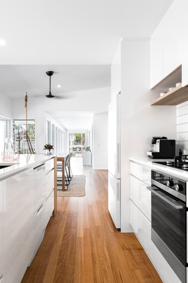 This is an example of a mid-sized modern galley open plan kitchen in Gold Coast - Tweed with a drop-in sink, flat-panel cabinets, white cabinets, quartz benchtops, white splashback, ceramic splashback, white appliances, medium hardwood floors, with island and white benchtop.