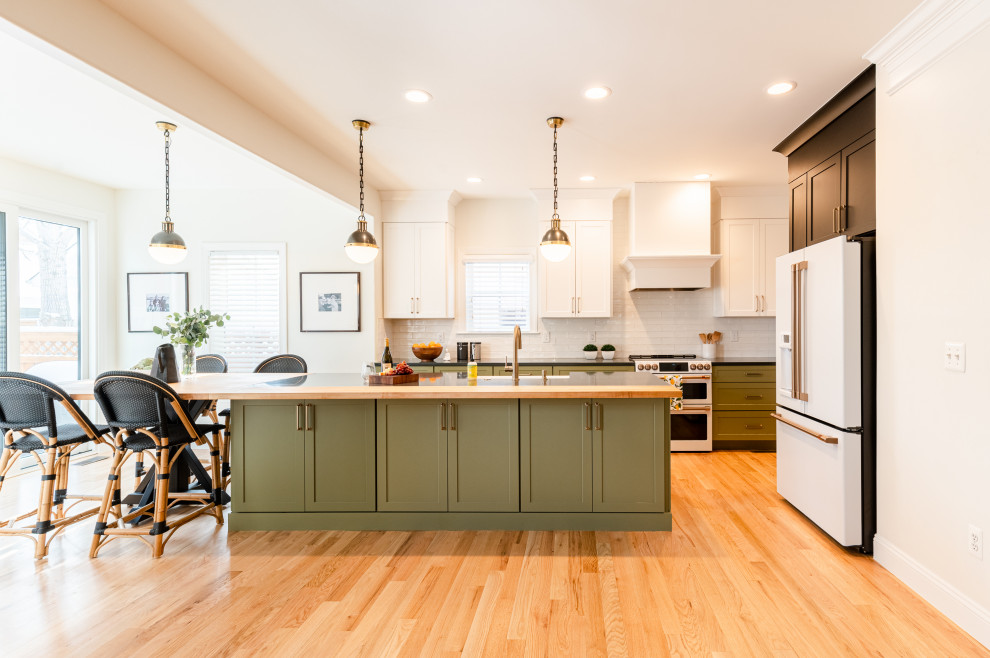 Large eclectic l-shaped eat-in kitchen in Denver with a farmhouse sink, shaker cabinets, green cabinets, wood benchtops, white splashback, ceramic splashback, white appliances, light hardwood floors, with island and black benchtop.