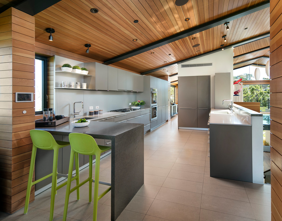 Photo of a large midcentury kitchen in Los Angeles with an undermount sink, flat-panel cabinets, grey cabinets, quartz benchtops, grey splashback, panelled appliances, porcelain floors, a peninsula and beige floor.