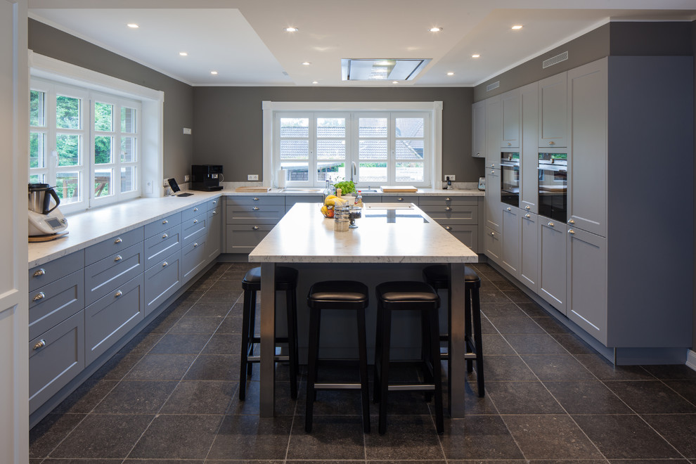 Photo of an expansive country u-shaped eat-in kitchen in Hamburg with a drop-in sink, beaded inset cabinets, grey cabinets, granite benchtops, grey splashback, black appliances, porcelain floors, with island, black floor and white benchtop.