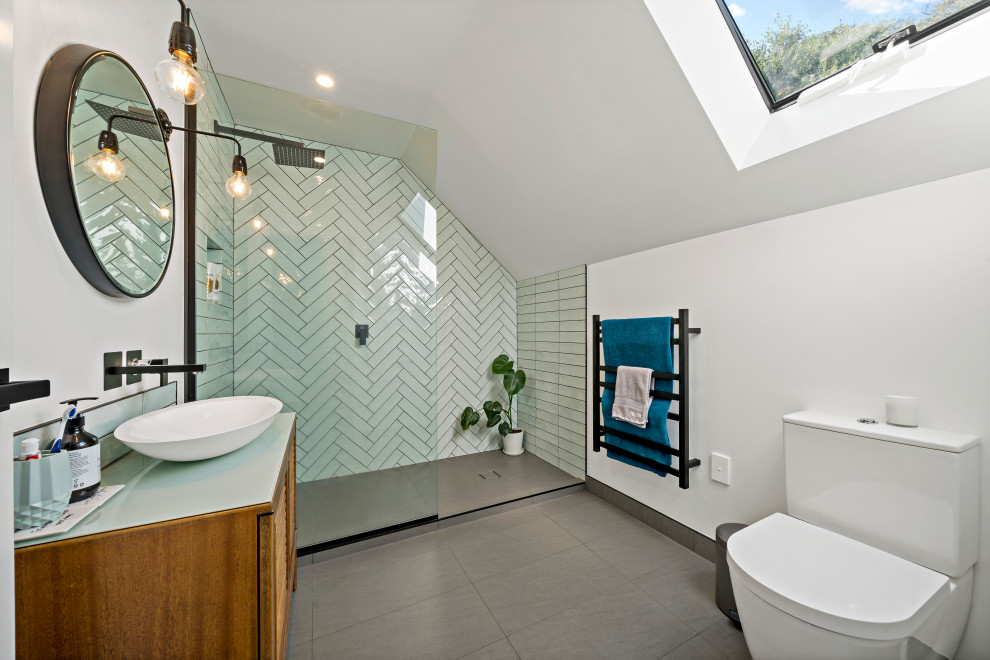 Mid-sized 3/4 bathroom in Auckland with furniture-like cabinets, brown cabinets, an open shower, a two-piece toilet, green tile, subway tile, white walls, porcelain floors, a vessel sink, glass benchtops, grey floor, an open shower, green benchtops, a single vanity, a freestanding vanity and vaulted.
